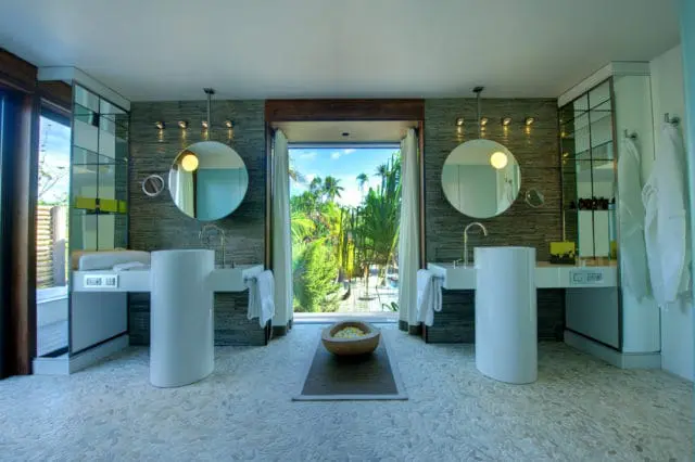 Modern bathroom with dual white pedestal sinks, round mirrors, and a large window overlooking a tropical garden.
