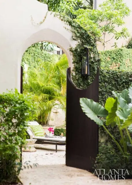 Arched entryway to a lush courtyard with palm trees, a chaise lounge with a green patterned pillow, and a dark wooden gate.