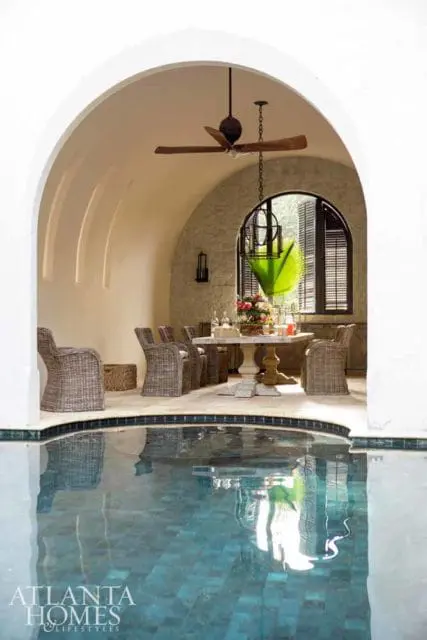 Covered patio with a marble-top dining table, wicker chairs, and a ceiling fan, overlooking a blue tiled pool.