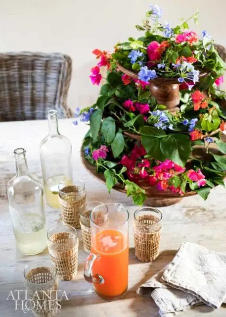 A wooden tiered stand overflowing with vibrant pink, red, and blue flowers and green leaves. On a light wood table are two glass bottles, a pitcher of orange juice, and several glasses with woven cozies.
