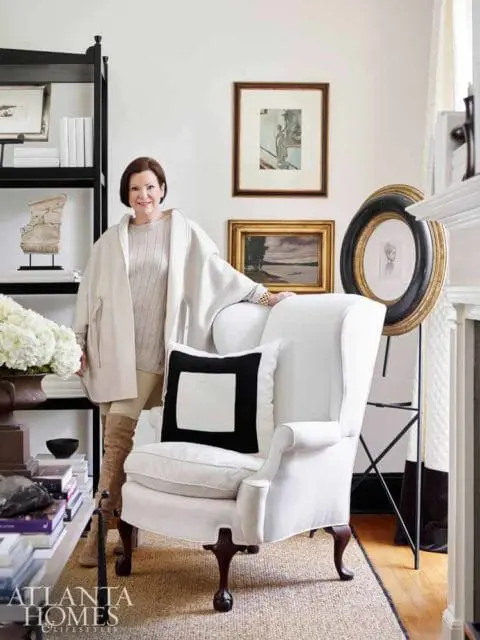 Woman in cream coat standing next to white wingback chair with black and white pillow. Black bookshelf with art and books is to the left.