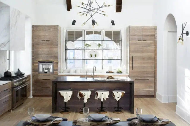 Modern kitchen with wood-paneled cabinets, a dark wood island with cowhide-covered stools, and a large window with black grid shelves displaying white dishes.