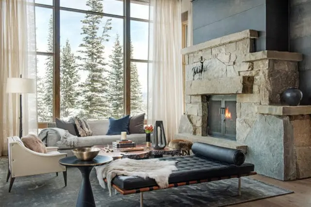 Living room with large stone fireplace, leather daybed, neutral sofa, and floor-to-ceiling windows overlooking a snowy forest.