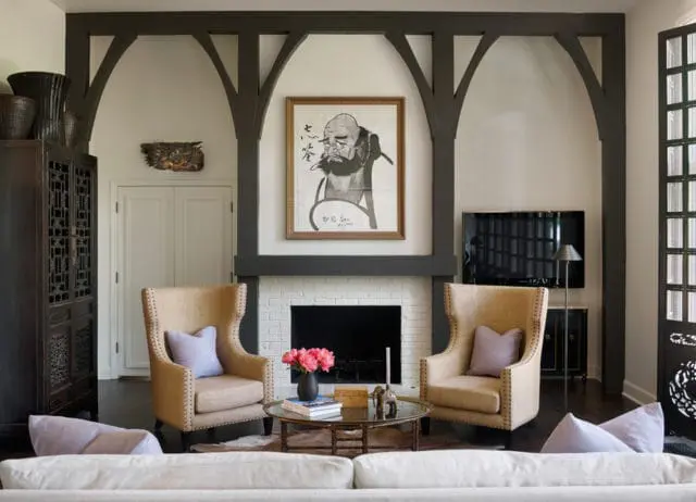 Living room with white brick fireplace, dark wood beams, two tan wingback chairs with lavender pillows, and a framed painting of a man above the mantel.