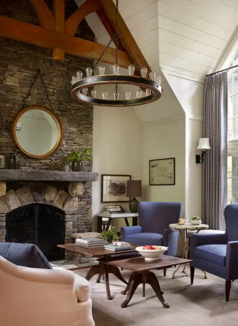 Living room with stone fireplace, vaulted wood-beamed ceiling, and blue upholstered armchairs. A circular chandelier hangs above two wooden coffee tables.