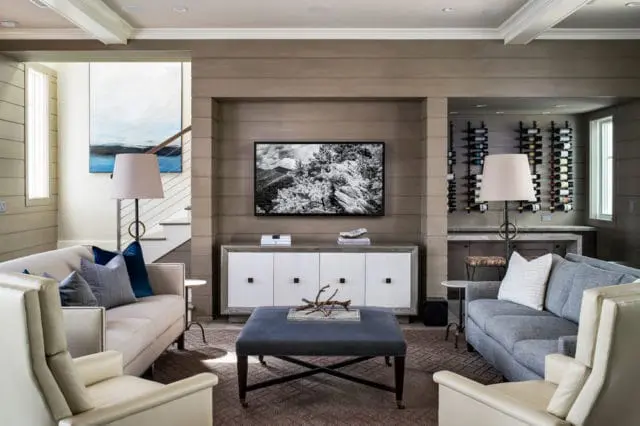 Living room with beige and blue sofas, cream armchairs, a dark blue ottoman coffee table, and a wall-mounted television above a white credenza. A wine rack is visible on the right.