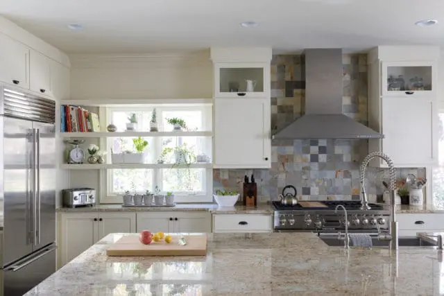 Cream kitchen with stainless steel refrigerator, open shelves displaying books and plants, a multi-colored stone tile backsplash, and a granite island countertop.