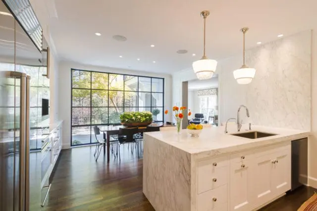 Modern kitchen with white marble island and countertops, stainless steel appliances, and a large grid window overlooking a garden.