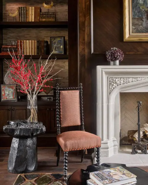 Living room with dark wood paneled walls, a stone fireplace, and bookshelves filled with books. A vintage chair with orange upholstery sits next to a black carved side table with a vase of red berries.