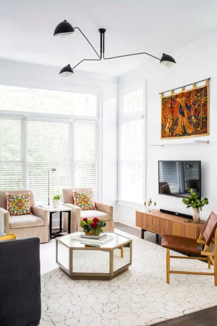 Living room with two upholstered chairs, a mirrored hexagonal coffee table, a wooden media console with a television, and a large tapestry hanging on the wall.