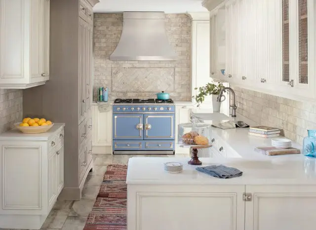 White kitchen with a large blue range, marble backsplash, and white countertops. A bowl of lemons sits on a counter to the left.