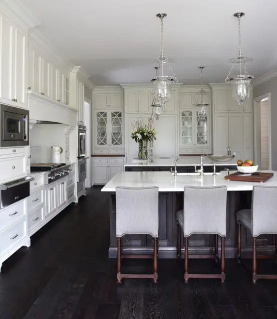 White kitchen with dark wood floors, marble island with gray upholstered stools, and custom cabinetry with glass-fronted upper cabinets.