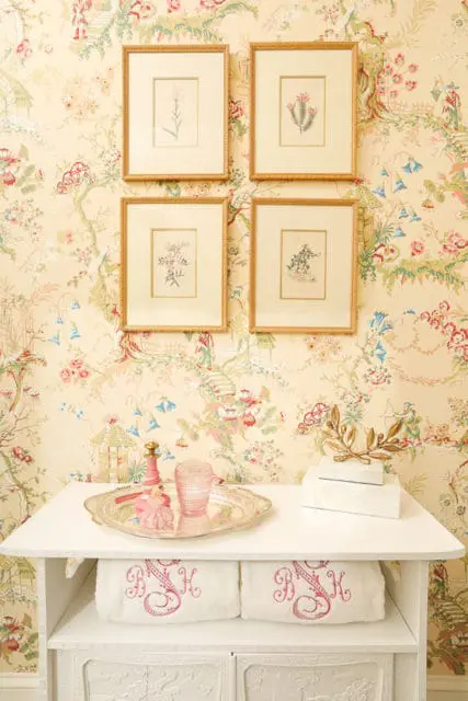 Powder room with chinoiserie wallpaper, four framed botanical prints, a white vanity with silver tray, pink glass, and monogrammed towels.