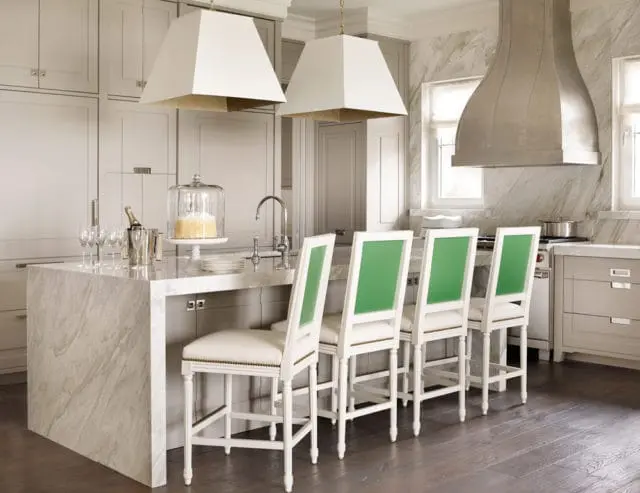 Kitchen island with marble countertop and white bar stools featuring green backs. White cabinetry and a stainless steel range hood are visible.