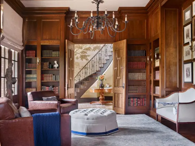 Library with dark wood paneling, built-in bookshelves filled with books, leather armchairs, a tufted ottoman, and a view of a staircase with tree-motif wallpaper.