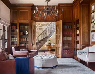Library with dark wood paneling, built-in bookshelves filled with books, a tufted ottoman, and a view of a staircase through double doors.