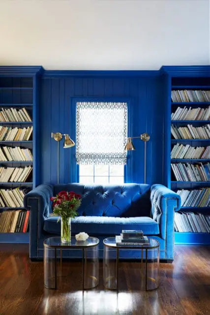 Blue tufted sofa in a room with floor-to-ceiling bookshelves, a patterned Roman shade window, and two glass and brass side tables.