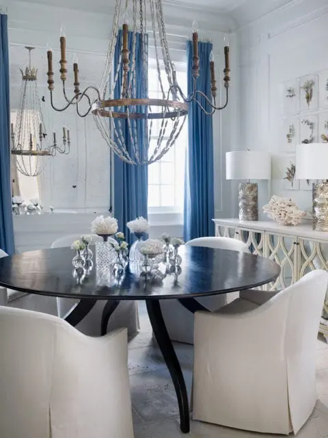 Round dark wood dining table with white slipcovered chairs, blue drapes, and ornate chandeliers in a room with mirrored walls and botanical prints.