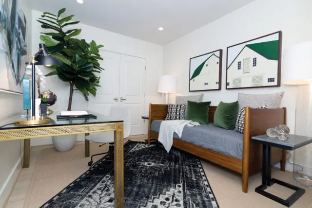 Home office with a glass-top desk featuring a gold frame, a rolling chair, a daybed with green and patterned pillows, and two framed artworks of barns on a white wall.