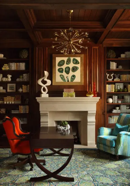 Library with dark wood paneling, stone fireplace, and built-in bookshelves. Features a gold sputnik chandelier, abstract art, an orange desk chair, and a striped armchair.