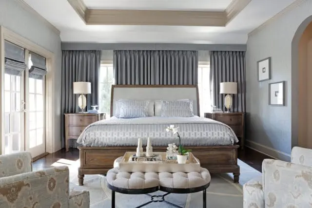 Bedroom with a king-size bed featuring a light gray upholstered headboard and blue patterned bedding. Dark wood nightstands flank the bed, each with a lamp. Gray curtains hang behind the bed, and French doors are visible to the left.