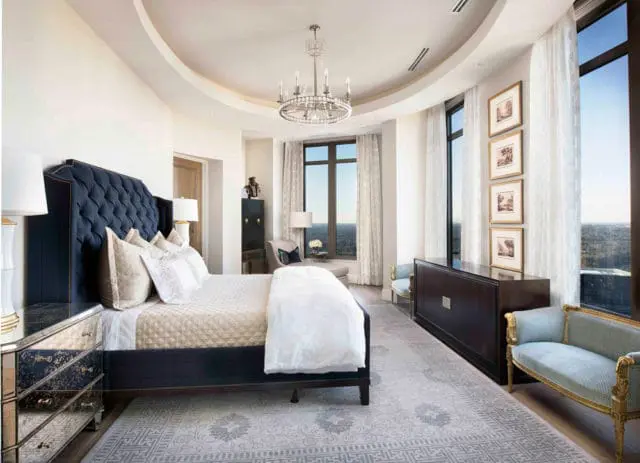 Bedroom with a navy tufted headboard, mirrored nightstand, patterned rug, and large windows with a city view.