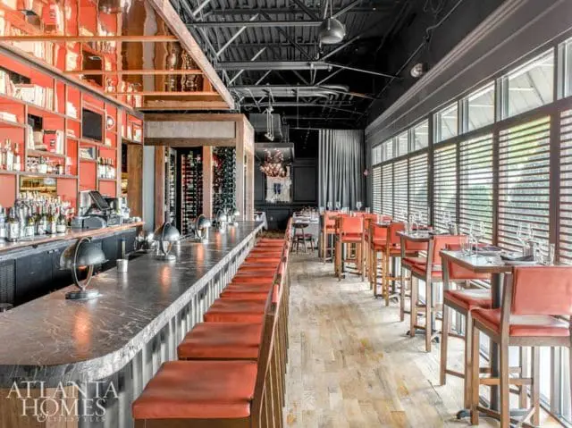 Restaurant interior with a long marble bar, orange leather bar stools, and high-top tables with orange chairs. Reflective copper ceiling and wood plank flooring.
