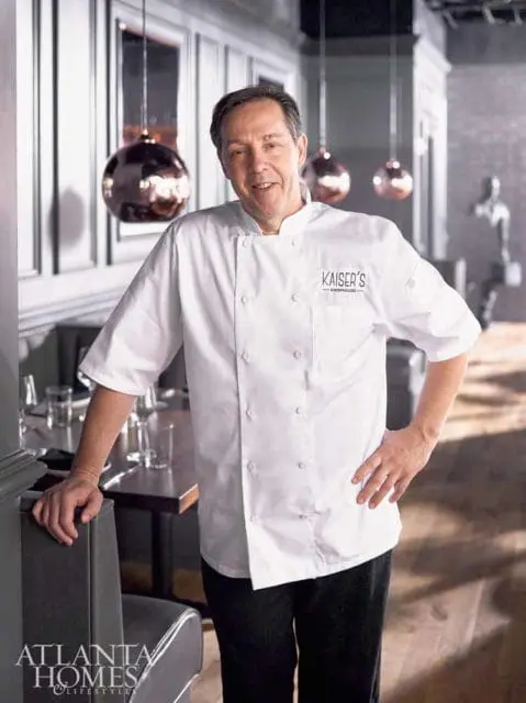 Chef in a white chef's coat with 'KAISER'S - CHOPHOUSE' logo, standing in a restaurant dining room with copper pendant lights.