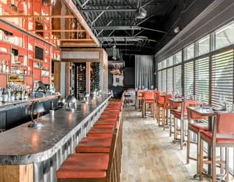 Restaurant interior with a long bar featuring red stools, a metallic backsplash, and dining tables with red chairs by large windows.