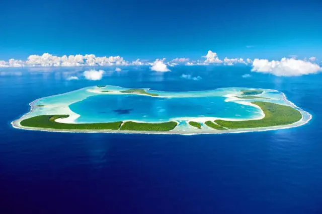 Aerial view of a lush green island surrounded by turquoise water and a deep blue ocean, with white clouds scattered across a bright blue sky.