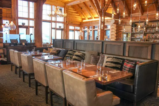 Log cabin interior dining area with leather chairs, plush upholstered booths, wooden tables set with glasses, and a bar in the background.