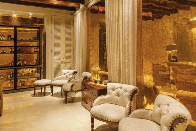 Lobby with tufted cream armchairs, a dark wood chest with tea set, and a display cabinet filled with bottles. Walls feature gold patterned panels and white slatted doors.