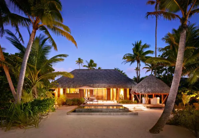 Small thatched-roof house with outdoor seating on a sandy beach at twilight.