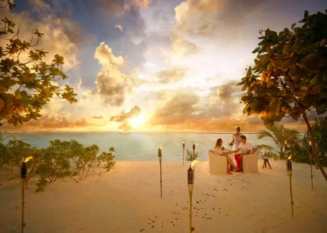 Couple enjoys a sunset dinner on a sandy beach with tiki torches.