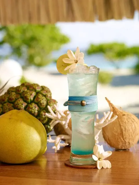 Blue cocktail with lime, flower, and pineapple on a wooden table.