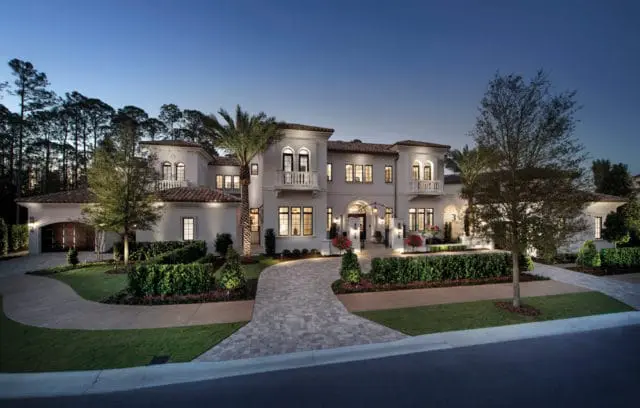 Mediterranean-style mansion with white stucco exterior, red tile roof, arched windows, and balconies, illuminated at dusk with manicured landscaping and a paver driveway.