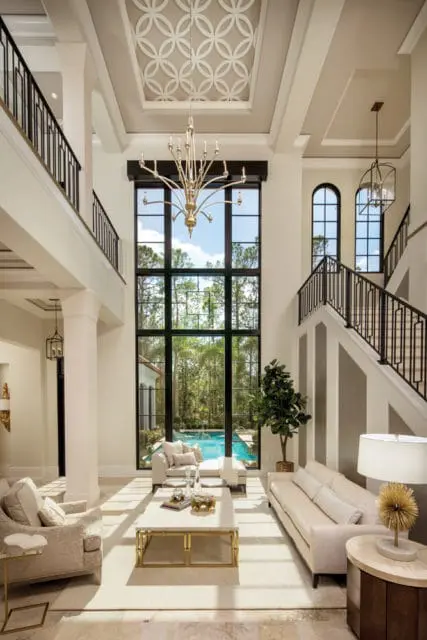 Living room with a two-story window overlooking a pool, featuring a gold chandelier, coffered ceiling with geometric pattern, neutral-toned furniture, and marble-topped coffee table.
