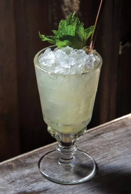 Close-up of a pale yellow cocktail in a stemmed glass filled with crushed ice and garnished with mint leaves and a copper straw.