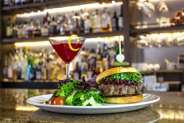 Gourmet burger with sprouts and cranberry sauce, served with a side salad and a red cocktail with a lemon twist, in front of a blurred bar background.