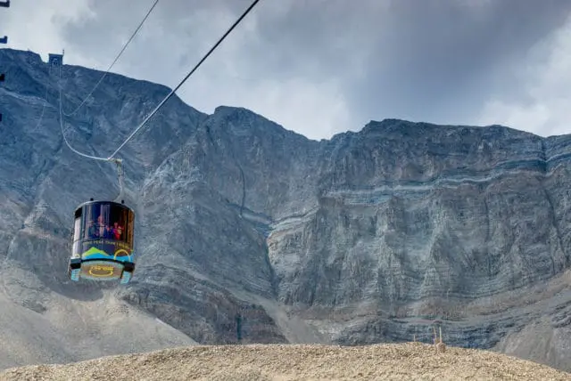 A tram car ascends a steep mountain under a cloudy sky, with layers of rock visible on the mountain face.