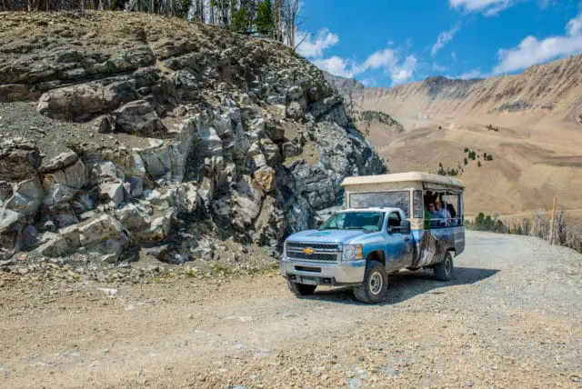 Light blue pickup truck with a tan pop-up canopy carrying passengers drives on a gravel road next to a rocky mountain slope.