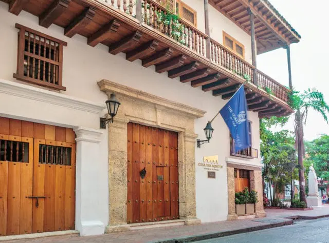 White building facade with large wooden doors, stone accents, a wooden balcony with ornate railings, and a blue flag displaying text.