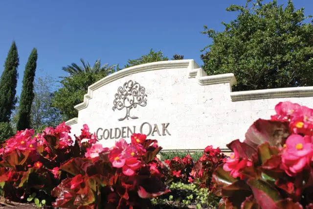 Stone monument sign with "GOLDEN OAK" in metallic lettering and a decorative tree emblem, surrounded by bright pink flowers and lush green trees under a clear blue sky.