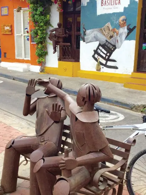 Rusty metal sculptures of people sitting on a bench in front of a building with a mural depicting a man reading a newspaper and text that reads "BASILICA Pizzeria, Cafe".