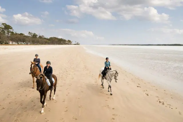 Three people horseback riding on a wide sandy beach with the ocean to the right and trees and houses in the distance to the left.