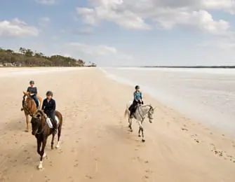 Three people on horseback ride along a wide sandy beach next to a calm body of water under a partly cloudy sky.