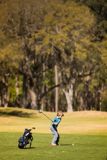Young boy in a teal shirt and grey pants swings a golf club on a sunny golf course, with a golf bag to his left.