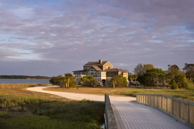 Large, multi-story house with gray siding and white trim, situated on a waterfront with a wooden boardwalk in the foreground and a winding path leading to the house.
