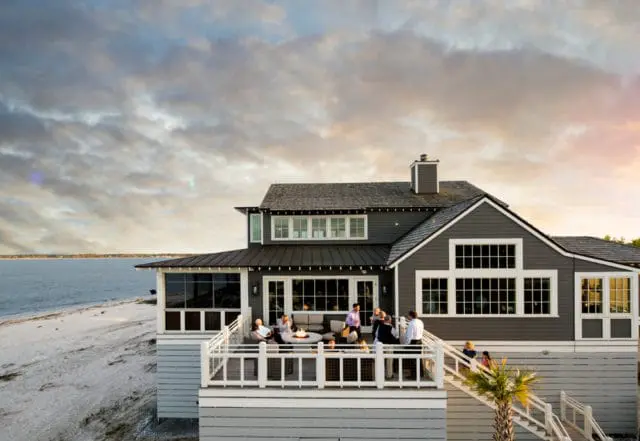 Dark gray shingle-sided house with white trim and multiple windows, featuring a large deck with people gathered around a fire pit overlooking a body of water and sandy beach.