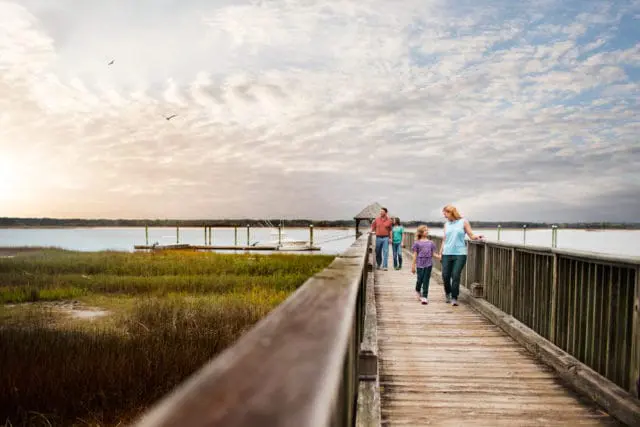 Family walking on a wooden pier over marshland with a calm bay and cloudy sky